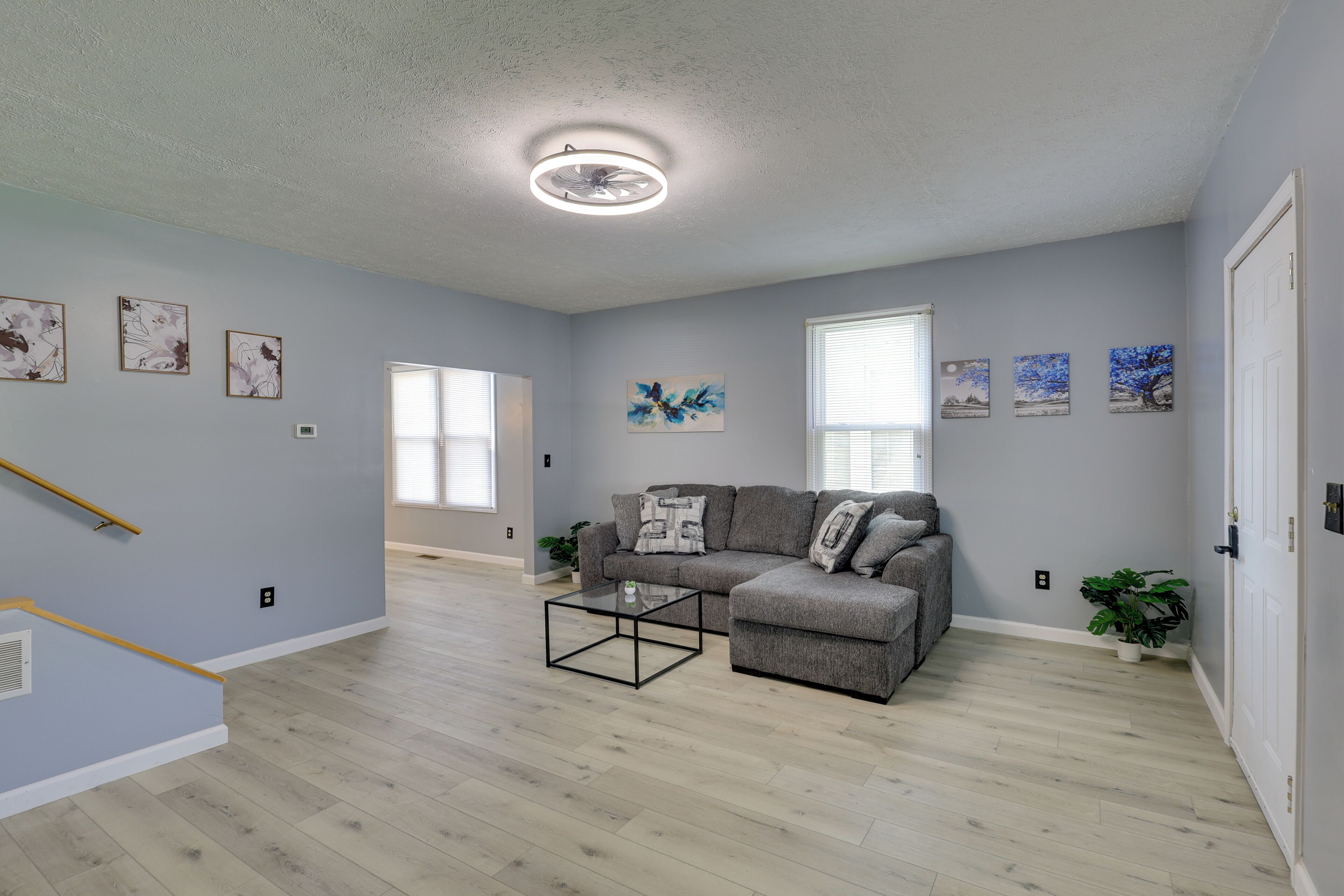 A spacious living room with a grey sofa, a glass coffee table, and artwork on the walls.