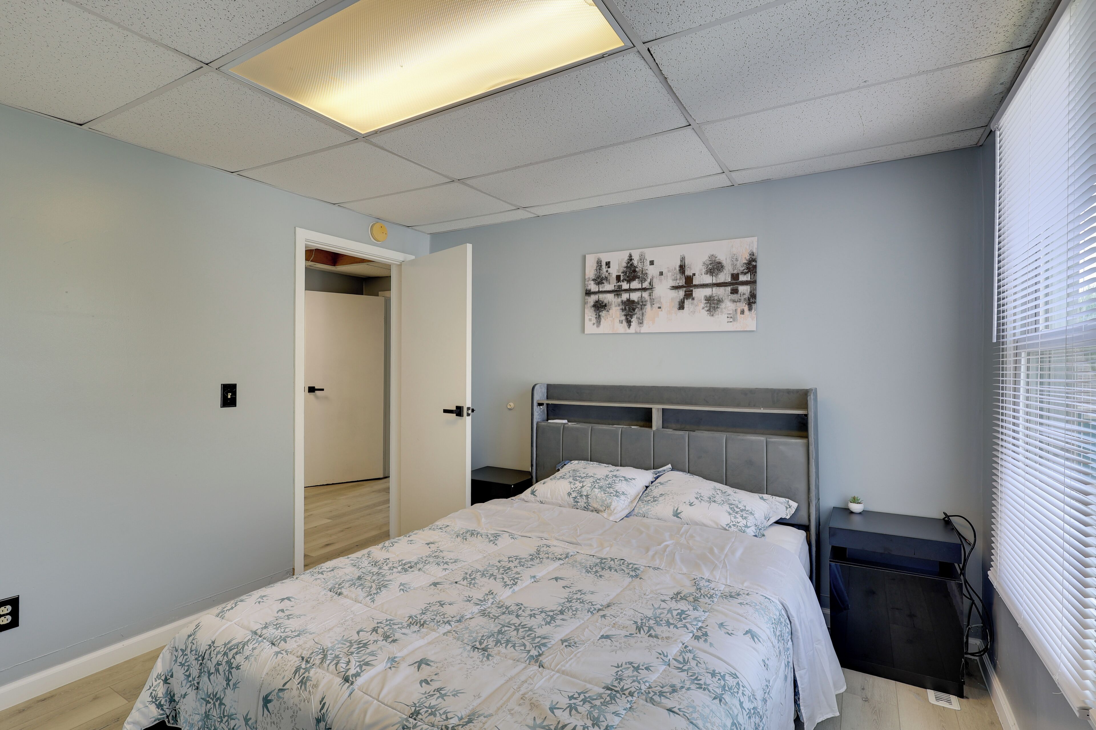 Un dormitorio con cama, mesita de noche, una obra de arte en la pared, una puerta que da a otra habitación y una lámpara en el techo.