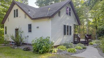 Une maison à deux étages avec un porche à l’avant et une terrasse, entourée d’un jardin et d’arbres.