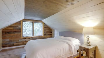 Une chambre avec un plafond en pente, un mur en bois, une fenêtre, un lit et une table de chevet avec une lampe.