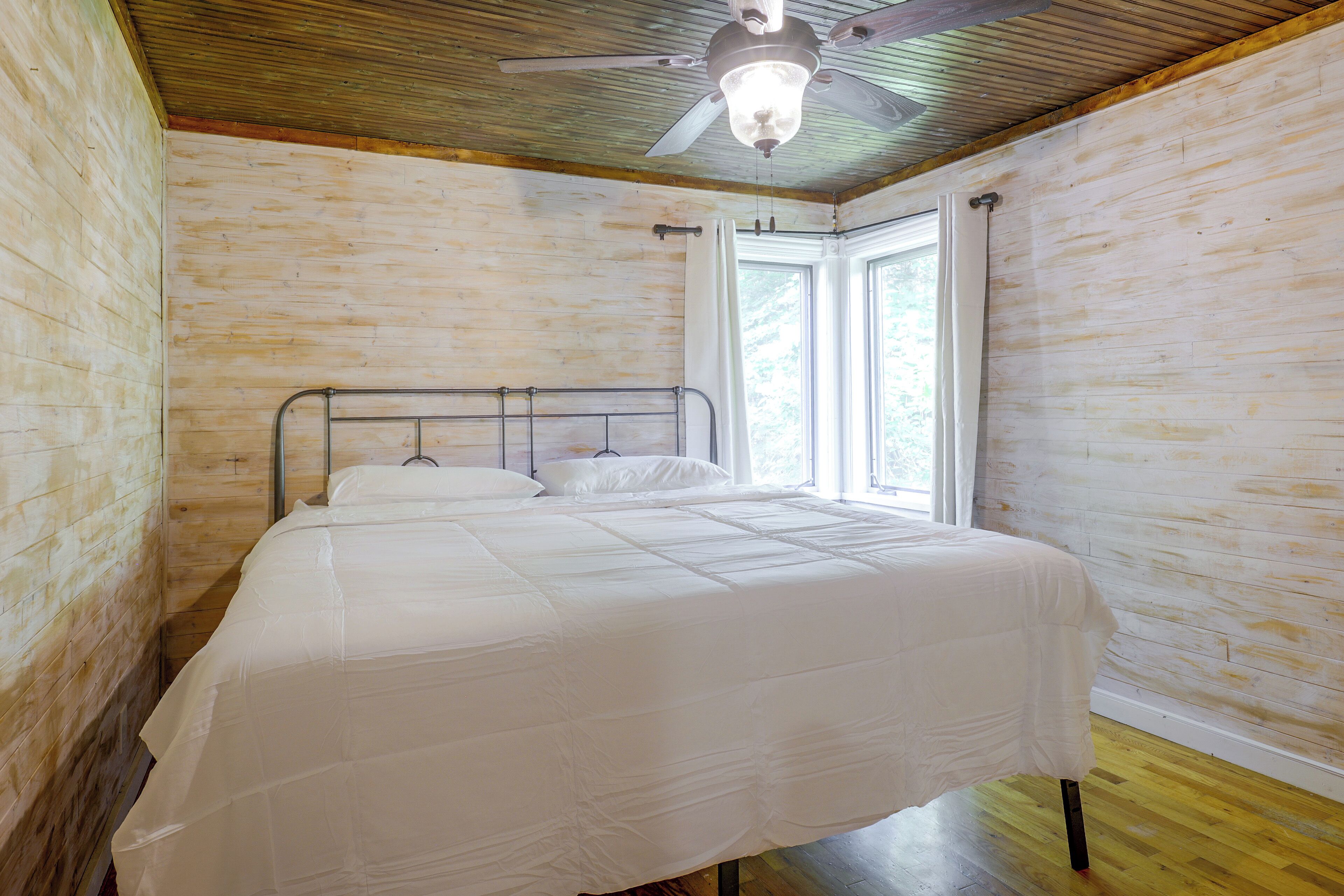 A bedroom with a bed, wooden walls, a ceiling fan, and a window.
