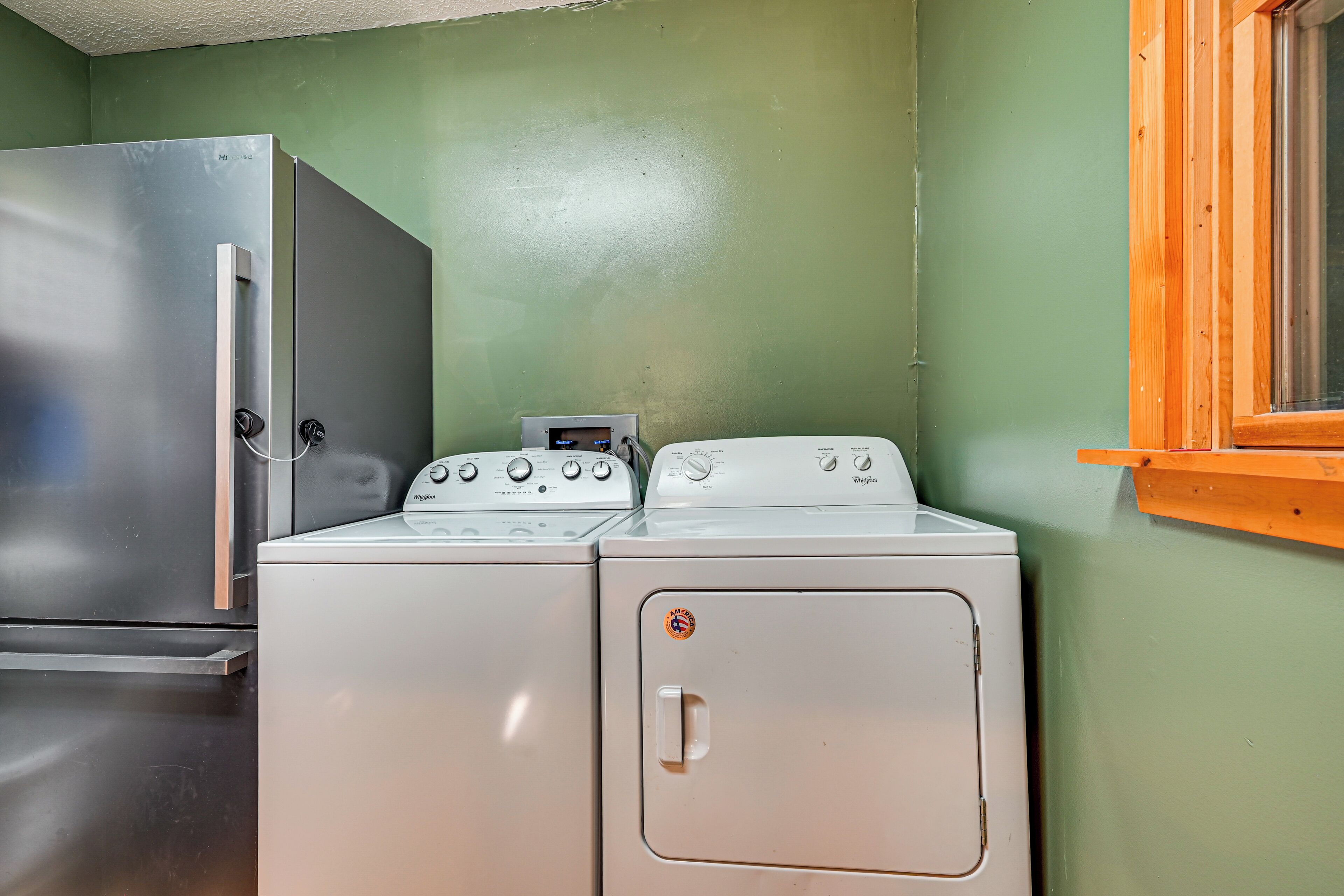Une petite buanderie équipée d’un réfrigérateur en acier inoxydable, d’un lave-linge et d’un sèche-linge blancs, et d’une fenêtre en bois.