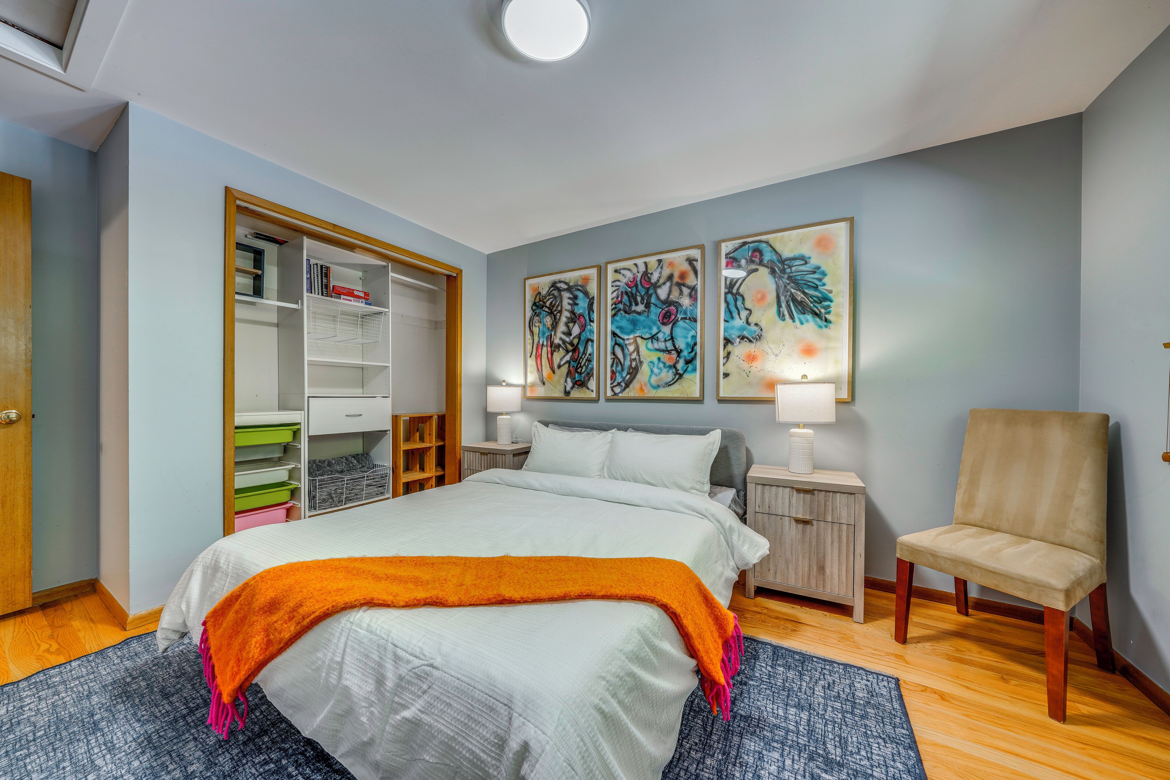 A bedroom with a bed, a chair, a nightstand, a lamp, a wardrobe, and artwork on the wall.