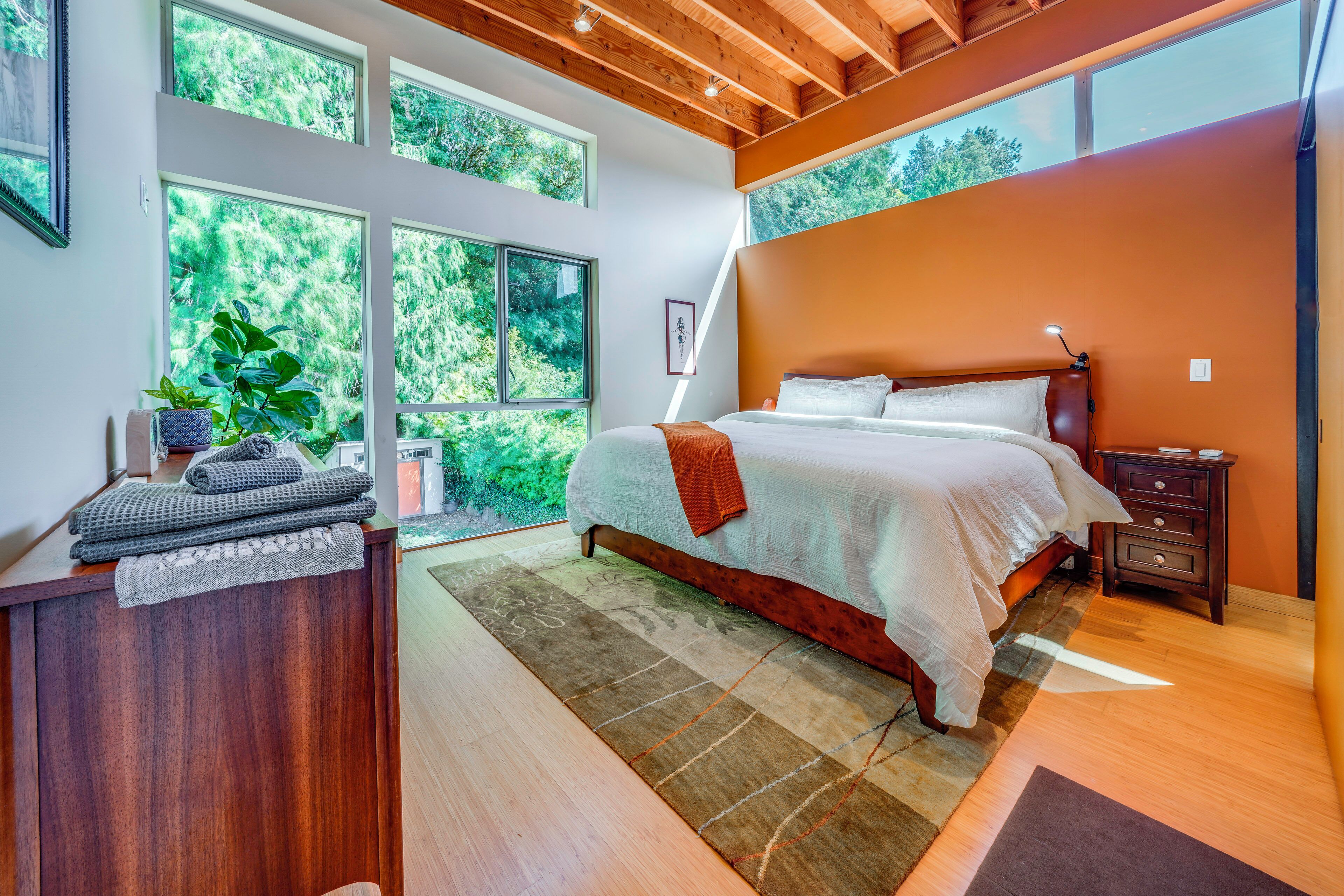 A bedroom with a large bed, a desk, and a view of greenery through the windows.