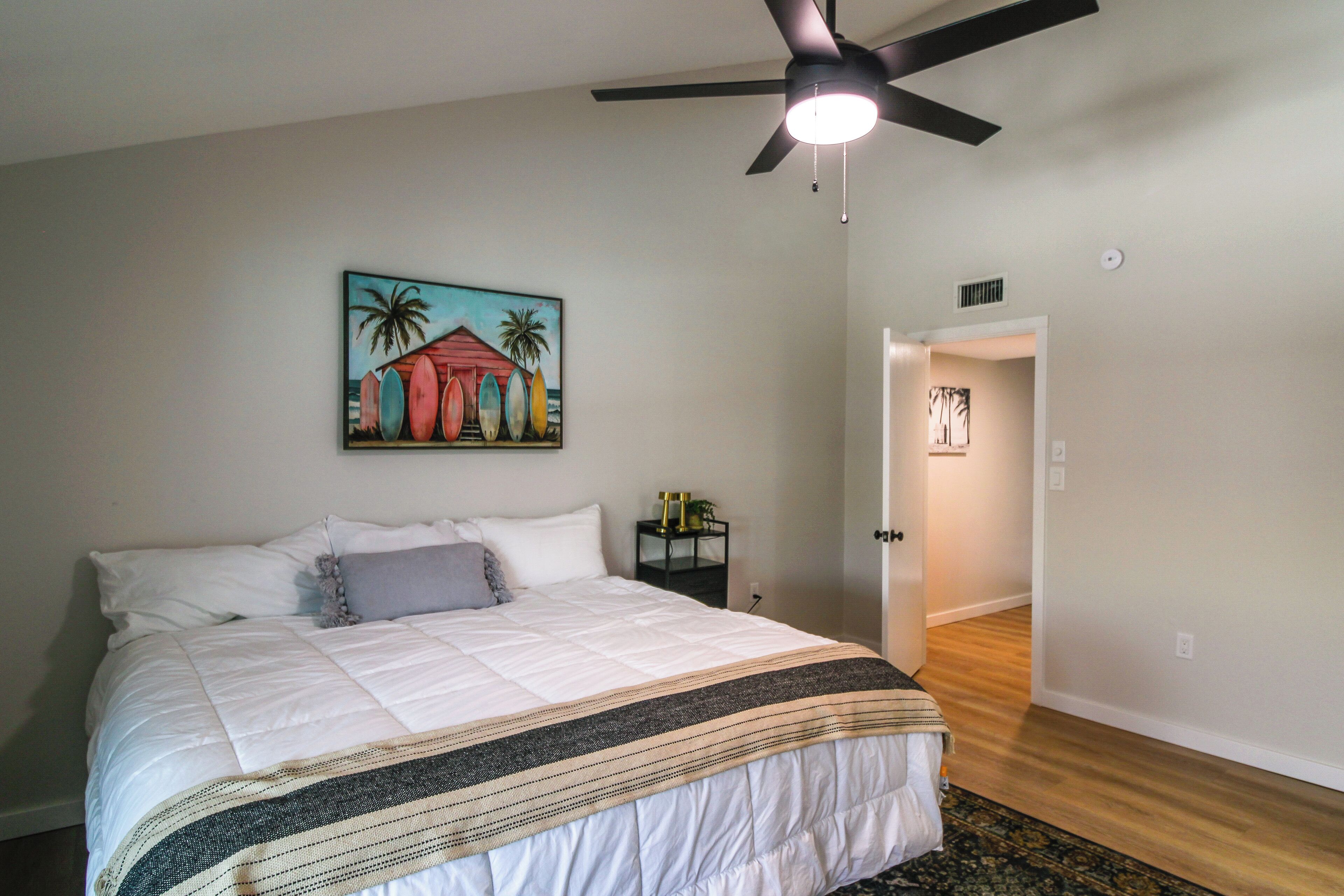 A bedroom with a bed, a ceiling fan, a painting, and a hallway.