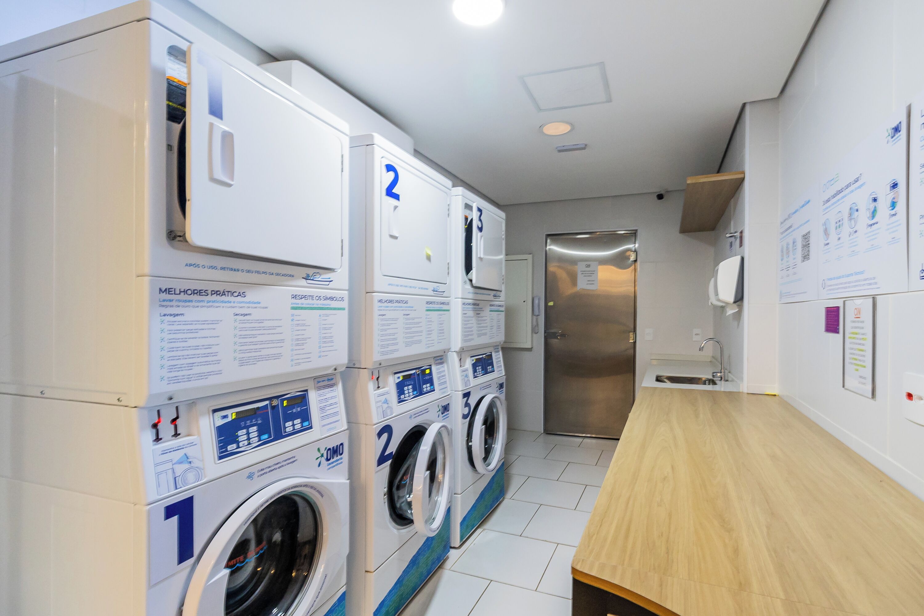 A laundry room with multiple washing machines, a sink, and informational posters on the wall.