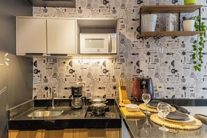 A kitchen with a microwave, coffee maker, and a modern stove. There is a wooden shelf above the counter holding cups and a glass. - Moema | Studio + 9 min Airport and Parking (São Paulo)