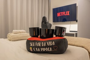 A hotel room with a bed, a pillow with text, a TV displaying 'NETFLIX', and a KFC cup. - Moema | Studio + 9 min Airport and Parking (São Paulo)