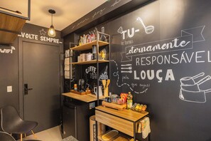 A modern kitchen with a chalkboard wall displaying text and drawings, wooden shelves with kitchenware, a coffee machine, and a small wooden table with various items. - Moema | Studio + 9 min Airport and Parking (São Paulo)