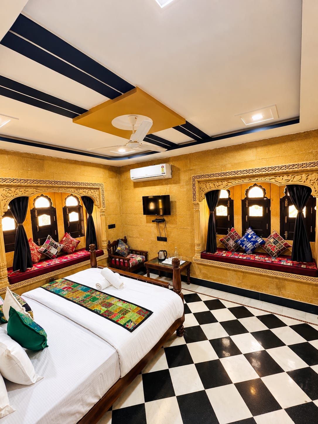 Una habitación de hotel con piso a cuadros, una cama con colcha de colores, una zona de estar con cojines y un televisor.