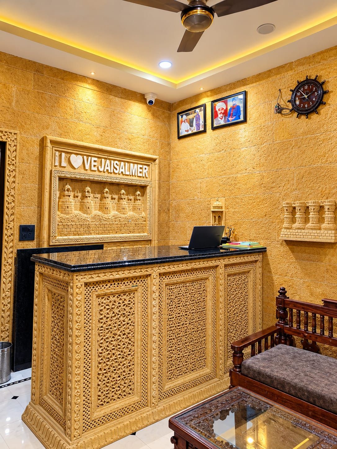 A reception desk with intricate carvings, a ceiling fan, a clock, and framed pictures on the wall.