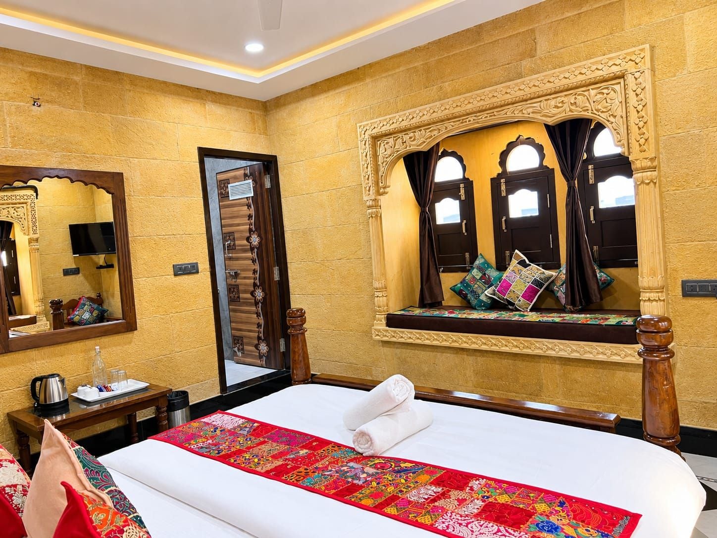 Habitación de hotel con una cama grande, una mesita de noche con hervidor, un espejo y una ventana con cortinas.