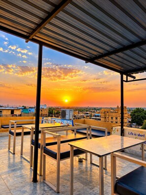 Un área de estar al aire libre con mesas y sillas, con vista a un atardecer y edificios en el fondo.