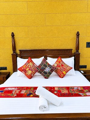 Una cama con cabecera de madera, dos almohadas rojas, una almohada estampada y una toalla blanca enrollada sobre una colcha con estampado rojo y blanco.