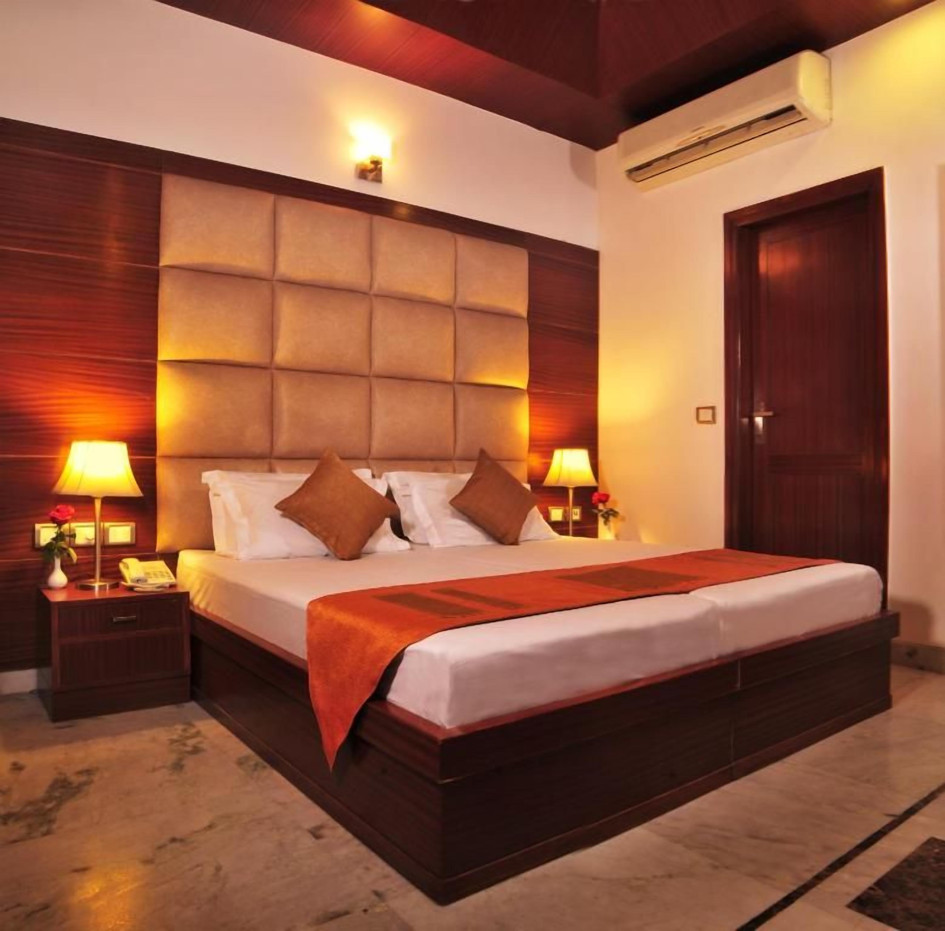 A hotel room with a large bed, two bedside tables with lamps, a wooden headboard, and an air conditioning unit.