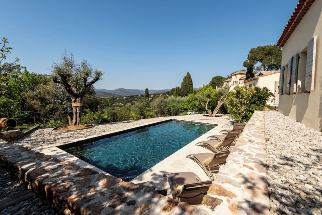 A rectangular swimming pool with a stone border, surrounded by lounge chairs and a pebble path.