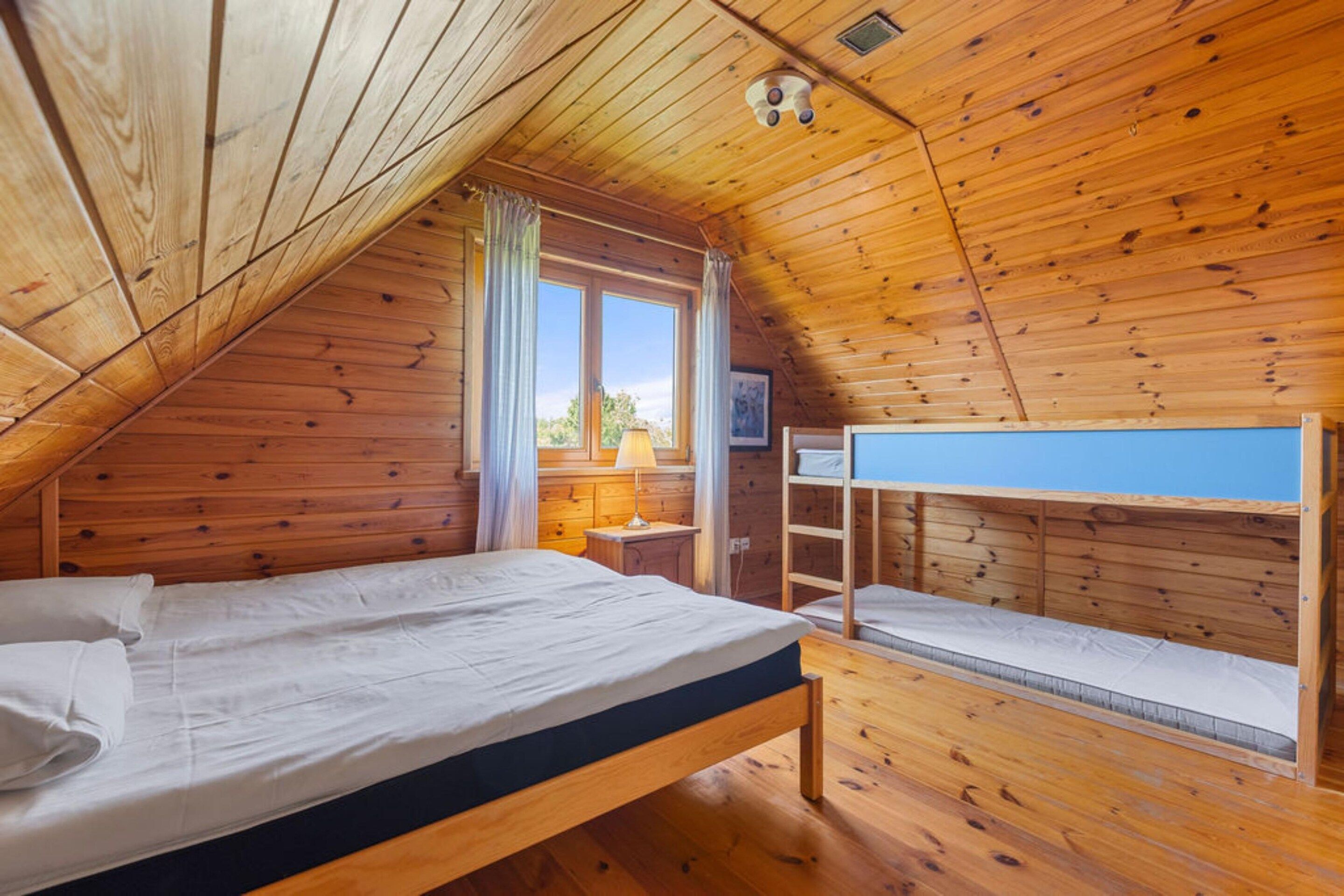 A room with a sloped ceiling, wooden walls, a bunk bed, a single bed, a window with curtains, and a lamp.