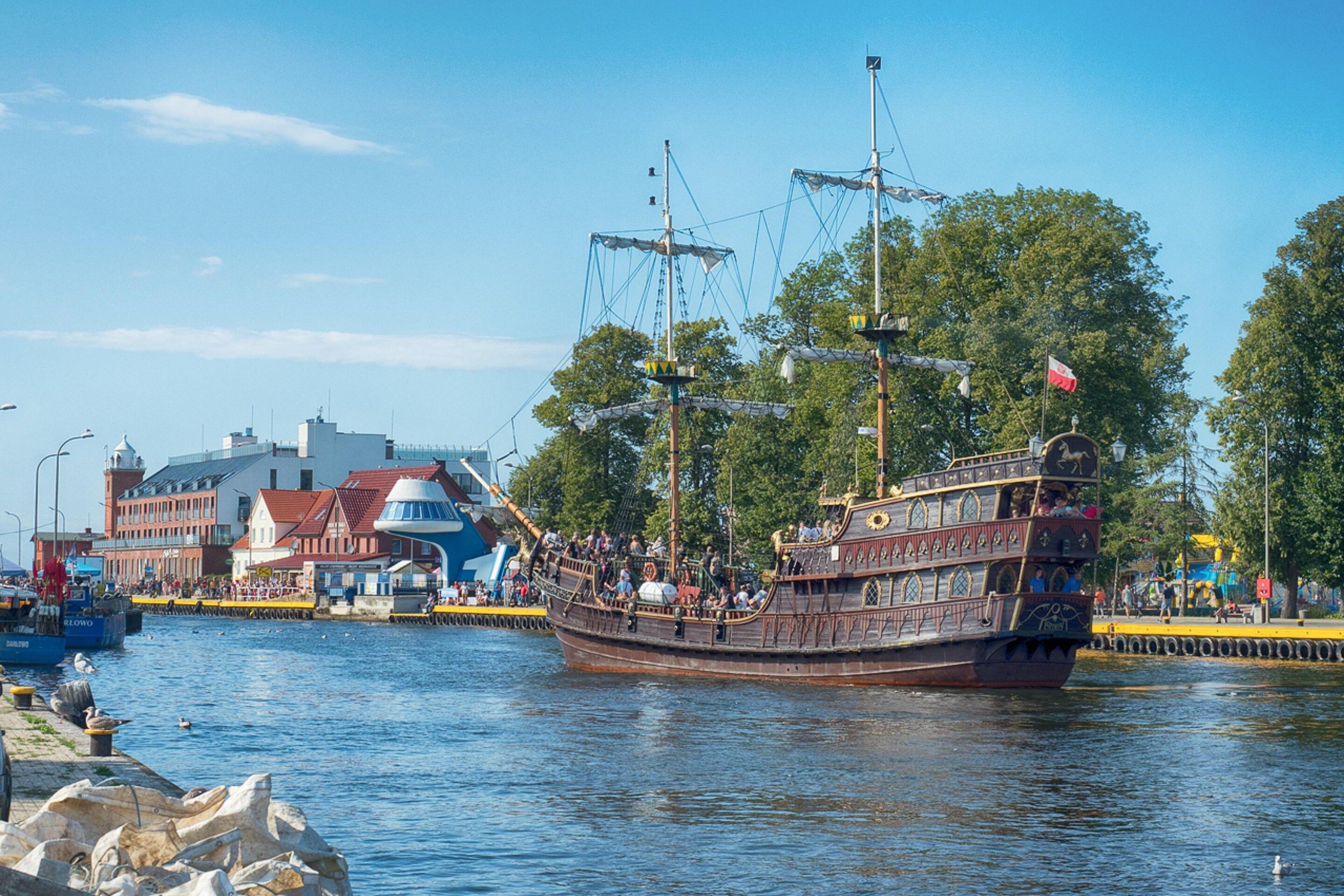 A historic sailing ship docked at a waterfront with buildings and trees in the background.