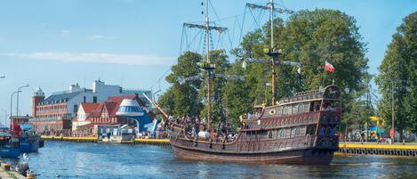 A historic sailing ship docked at a waterfront with buildings and trees in the background.