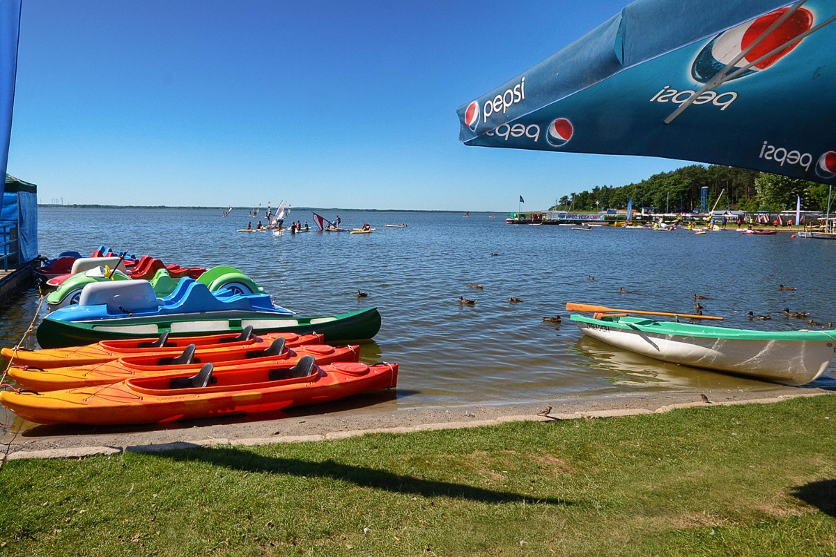 Blick auf einen See mit bunten Kajaks im Hafen, einem Pepsi-Sonnenschirm und blauem Himmel.