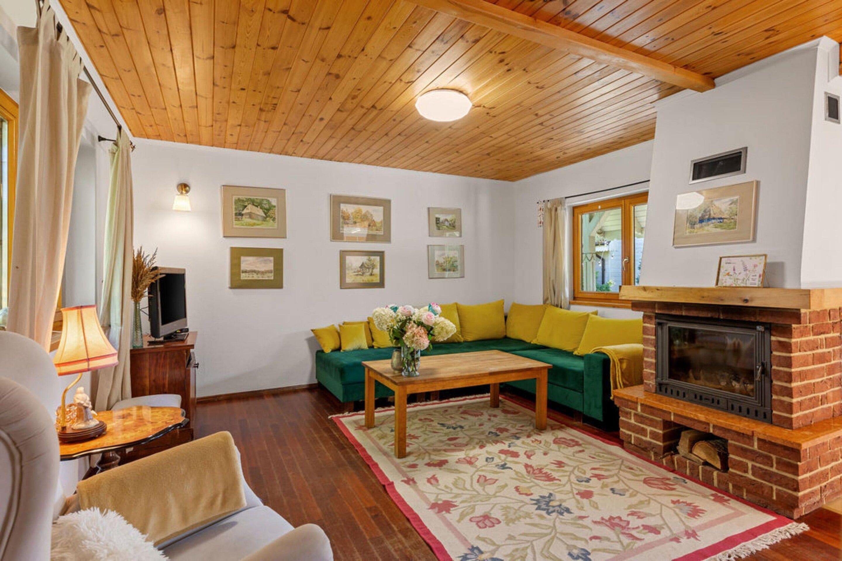 A cozy living room with a wooden ceiling, a green sofa, a fireplace, and framed pictures on the wall.