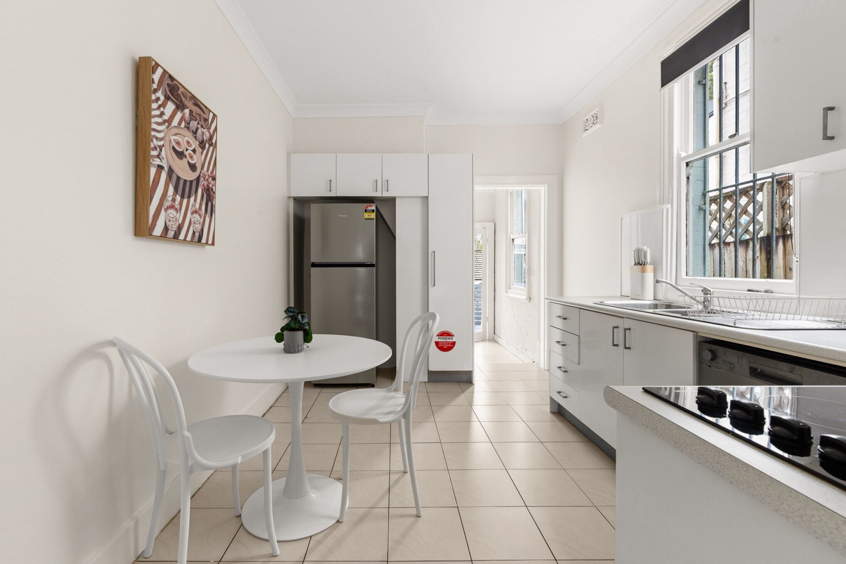 A modern kitchen with white cabinetry, a stainless steel refrigerator, a dining table with two chairs, and a window with a view of the outside.