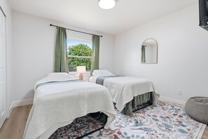 Quarto com duas camas de solteiro, janela com cortinas, abajur, espelho e tapete estampado.