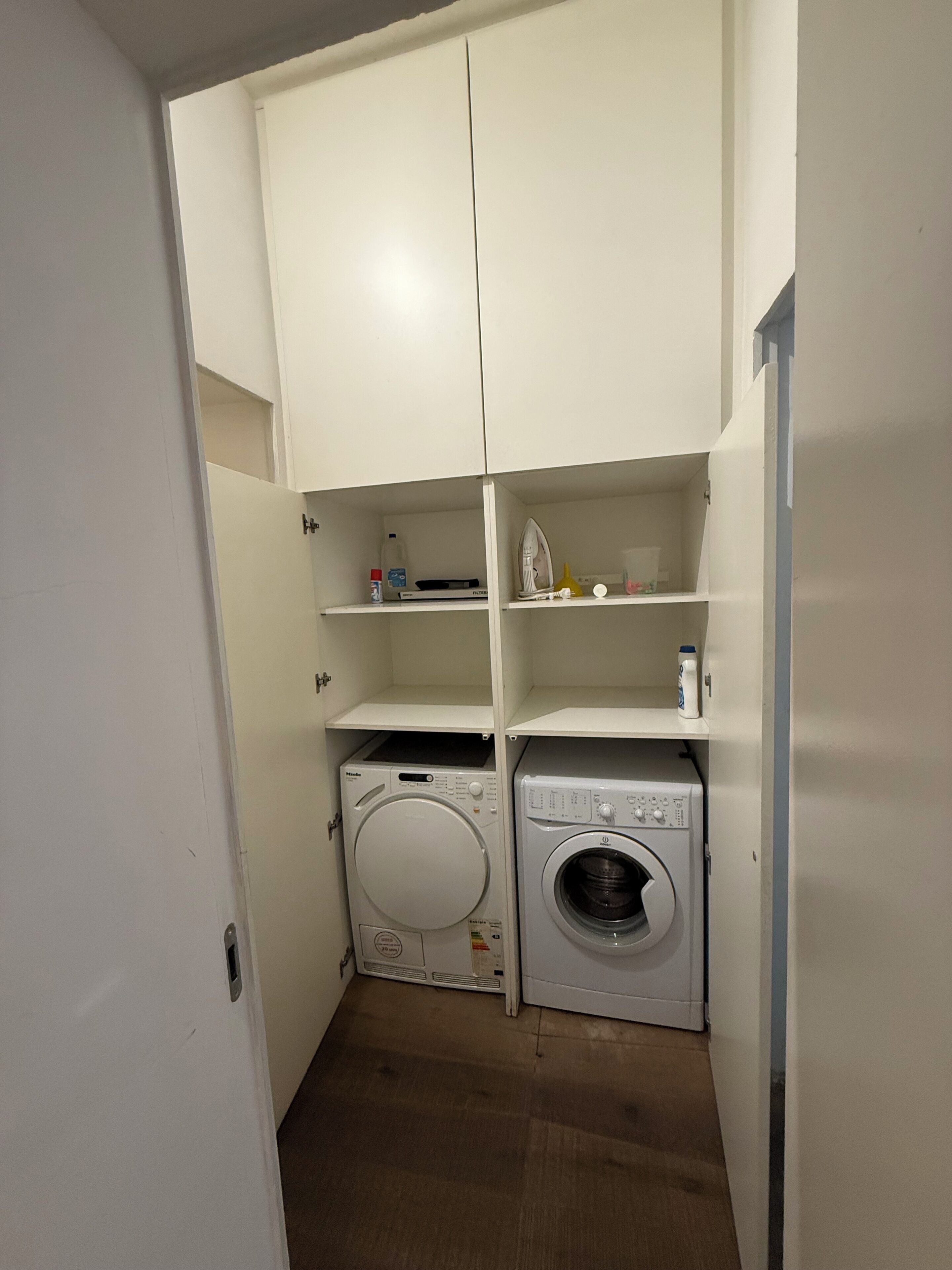Une buanderie équipée d’un lave-linge et d’un sèche-linge, d’étagères contenant des produits de nettoyage et d’une porte donnant sur une autre pièce.