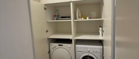A laundry room with a washing machine and dryer, shelves with cleaning supplies, and a door leading to another room.