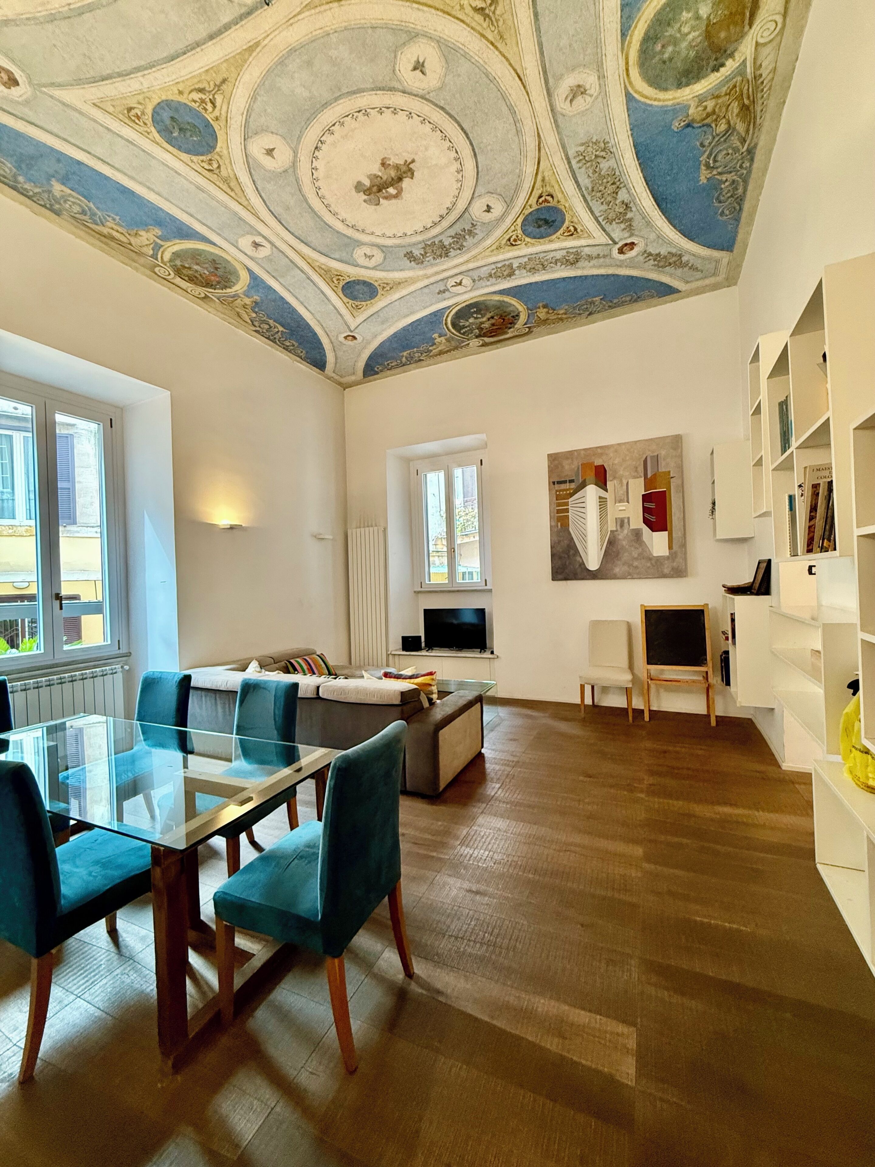 A spacious living room with a glass dining table, blue upholstered chairs, a sofa, a television, and a bookshelf.