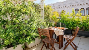 En udendørs terrasse med et bord dækket til to, omgivet af frodigt grønt og en hvid bygning i baggrunden.
