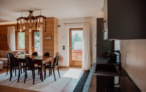 A modern kitchen with a built-in stove, a dining area with a wooden table and chairs, and a large window with curtains. - Dimora del Cervo | Luxury Dolomites Retreat. Spectacular mountain view (Falcade)