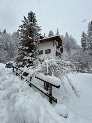 Ein mit Schnee bedecktes Blockhaus mit einer Holzterrasse und einem Baum vor der Tür, der ebenfalls mit Schnee bedeckt ist.