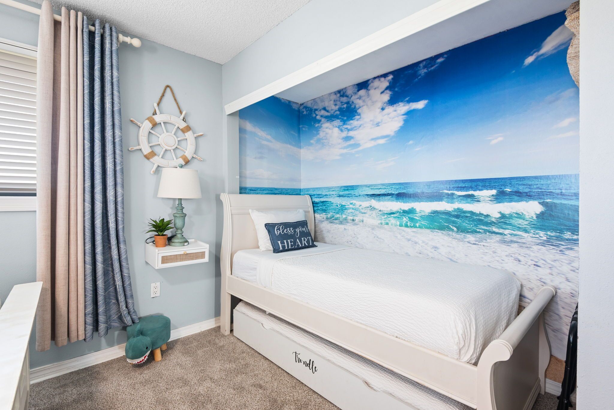 A bedroom with a bed, a wall mural of the ocean, a small shelf with a lamp and plant, and a decorative wall hanging.