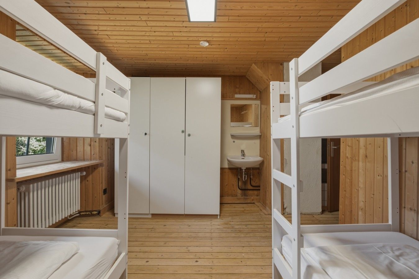 Quarto com beliche, piso de madeira, guarda-roupa branco e banheiro pequeno com pia.