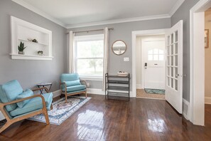 Een kamer met een houten vloer, een blauwe fauteuil, een kleine tafel, een plank met planten, een spiegel en een deur met een raam.