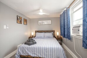 Ein Schlafzimmer mit Bett, Nachttischen, Deckenventilator, einem Fenster mit Vorhängen und Wandbildern.