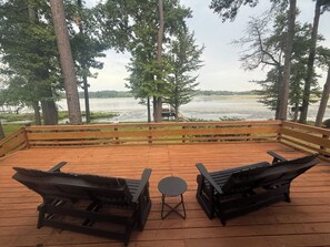 Eine Holzterrasse mit zwei Adirondack-Stühlen und einem kleinen Tisch, mit Blick auf einen See mit einem Steg und einem Boot.
