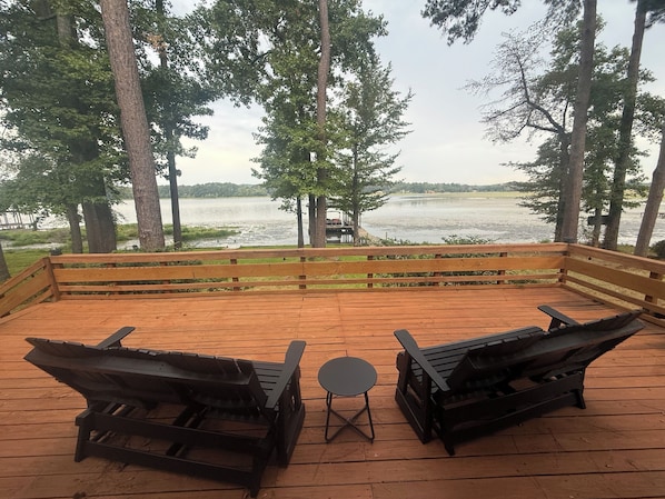 Um deck de madeira com duas cadeiras Adirondack e uma pequena mesa, com vista para um lago com um píer e um barco.