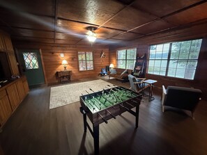 A cozy room with a foosball table, wooden walls, a ceiling fan, and large windows. - Charming 3-bedroom cabin with AC on Lone Star Lake (Lone Star)