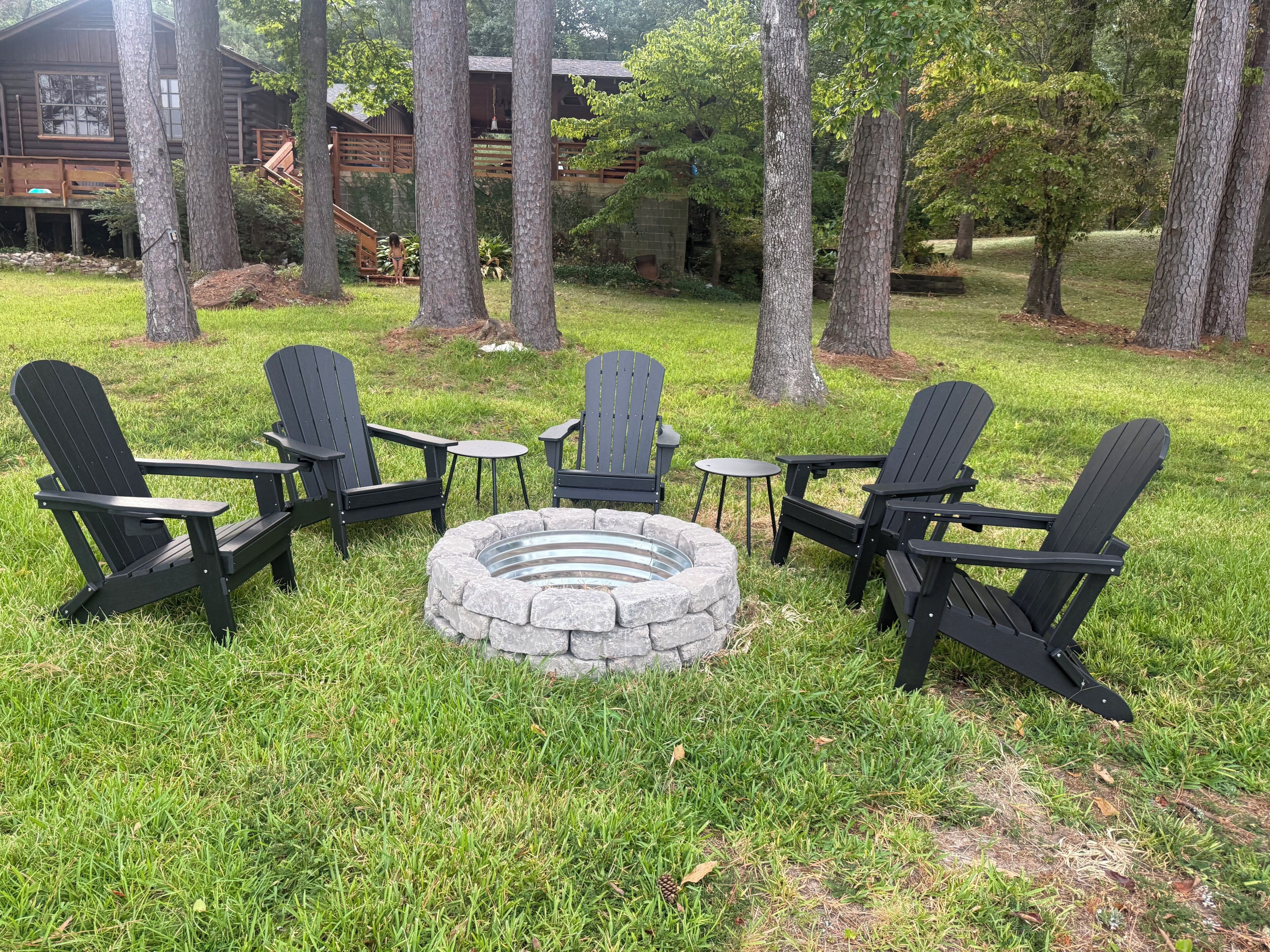 Een ronde vuurkuip van steen, omringd door vier Adirondack-stoelen, in een gazon met bomen en een houten gebouw op de achtergrond.