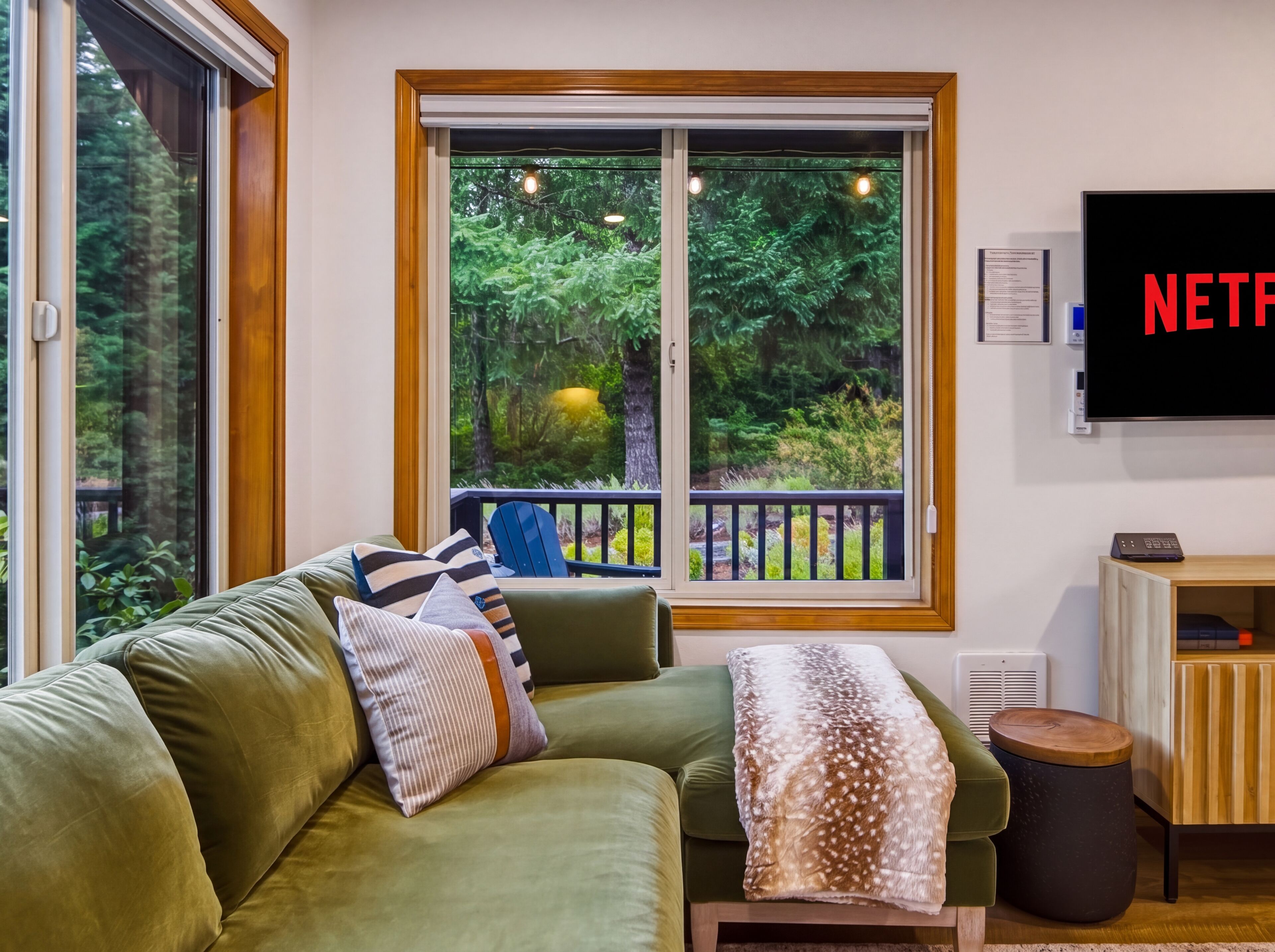 A living room with a green sofa, a large window with a view of trees, a flat-screen TV mounted on the wall, and a small wooden side table.