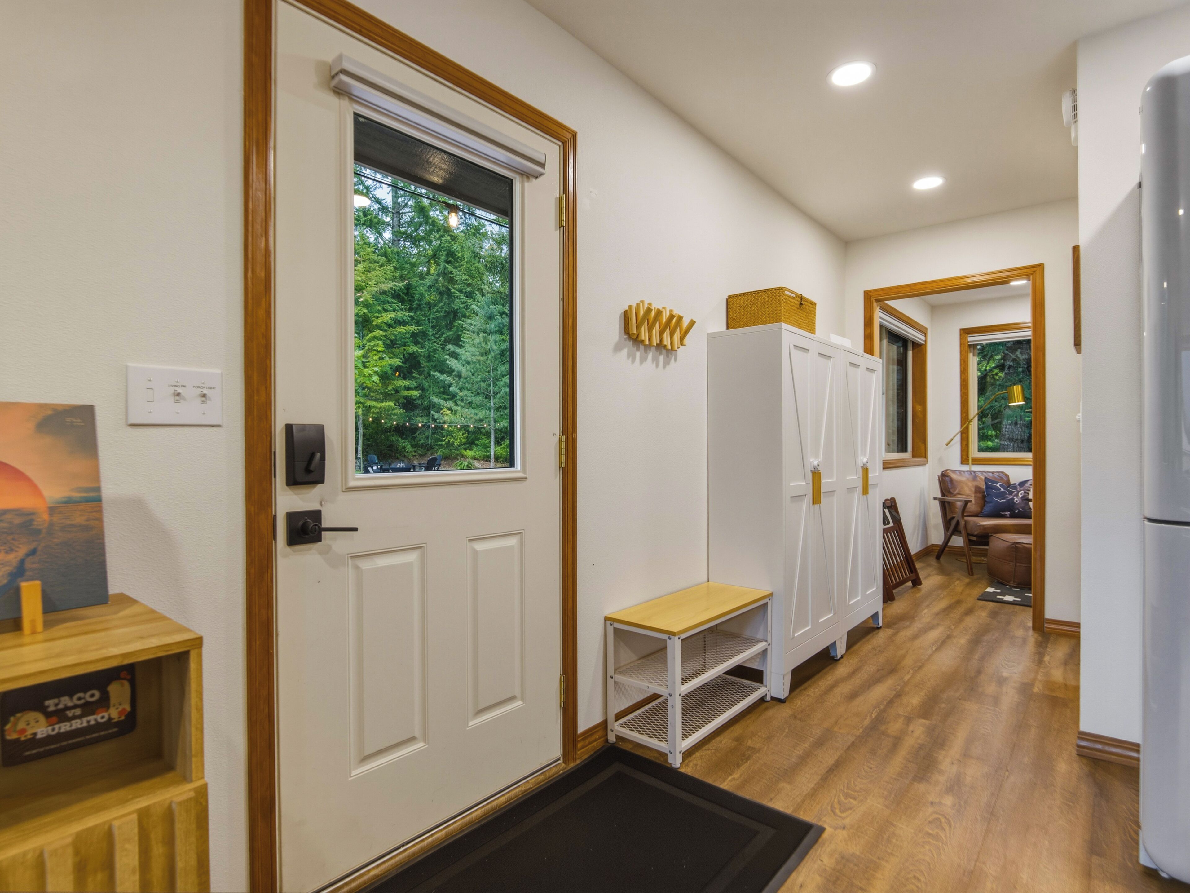 A hallway with a door leading to a room with a window overlooking a forest, a white cabinet, a small table, and a refrigerator.