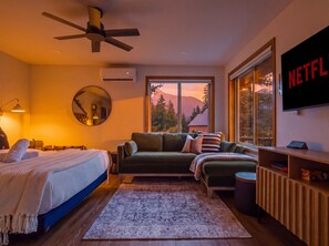 A modern bedroom with a bed, a sofa, a TV, and a large window with a view of mountains.