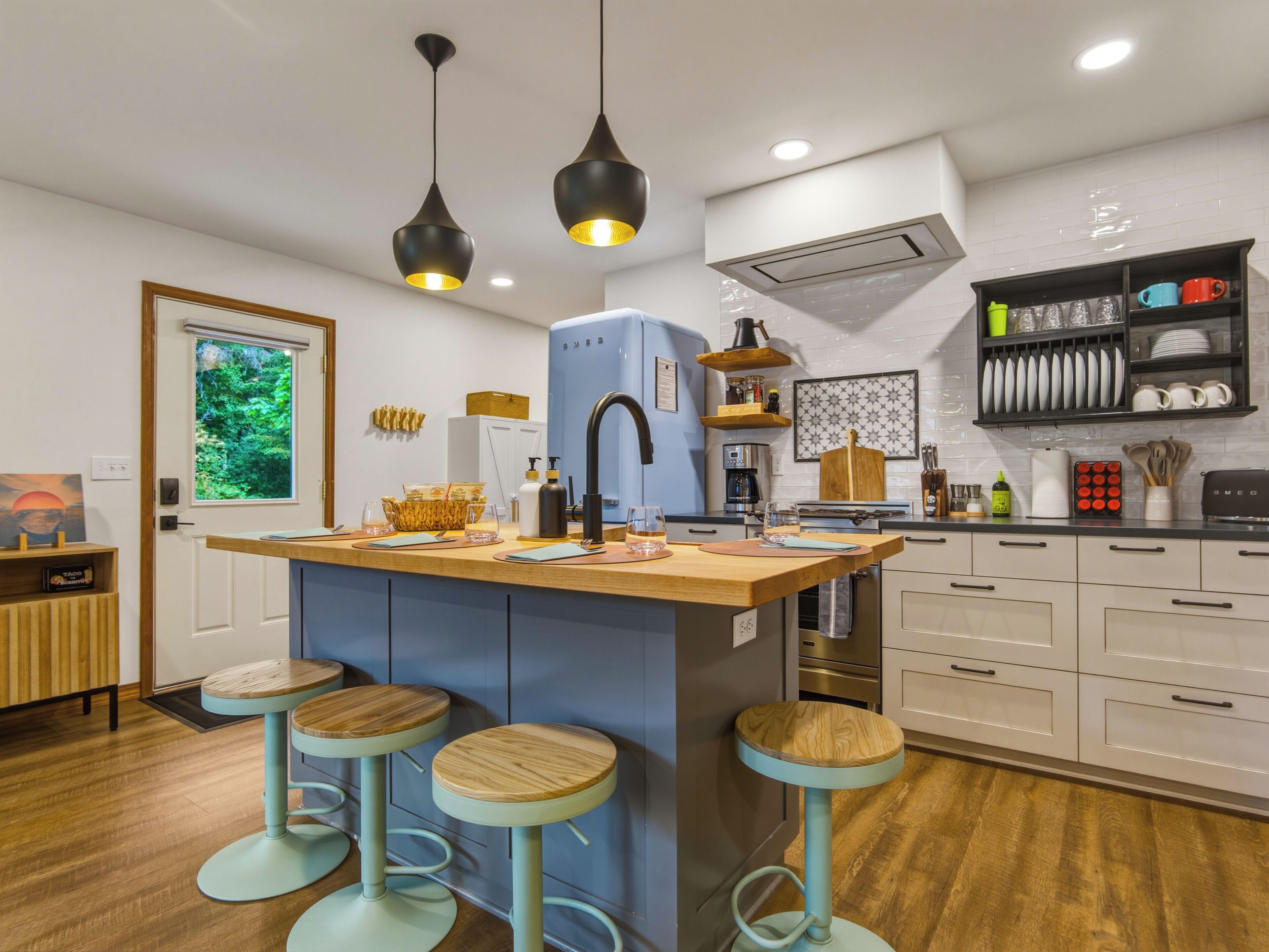 A modern kitchen with a central island, bar stools, pendant lighting, and a view of greenery outside.