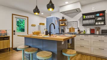 A modern kitchen with a central island, bar stools, pendant lighting, and a view of greenery outside.