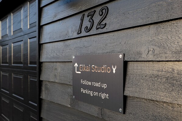 A wooden wall with a sign indicating the address '132' and directions to 'Elkai Studio' with an arrow pointing upwards and text stating 'Follow road up Parking on right.'