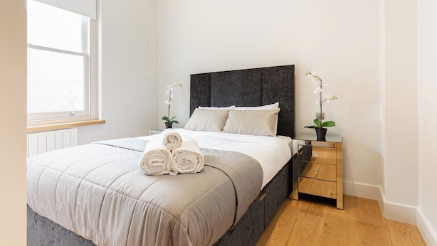 Un dormitorio con cama, mesita de noche, ventana y una orquídea en maceta.