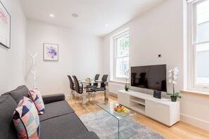 Ein modernes Wohnzimmer mit Sofa, Glastisch, Essbereich mit Stühlen, Flachbildfernseher und Fenster mit Tageslicht.