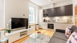 Una sala de estar moderna con un televisor de pantalla plana, una mesa de centro de vidrio, un sofá y una cocina con armarios oscuros y un microondas.